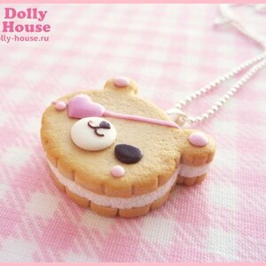 Puede incluir: Un collar de galleta con forma de oso rosa y blanco con una cadena plateada. La galleta tiene corazones rosas y una nariz negra.