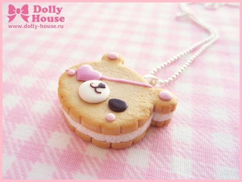 Puede incluir: Un collar de galleta con forma de oso rosa y blanco con una cadena plateada. La galleta tiene corazones rosas y una nariz negra.