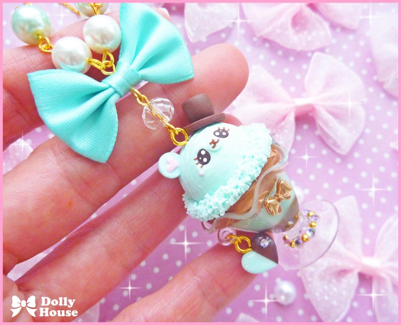May include: A light blue and white charm necklace with a green and brown ice cream cone charm. The charm features a cute cartoon face with black eyes and a brown hat. The necklace is made of beads and a gold chain.