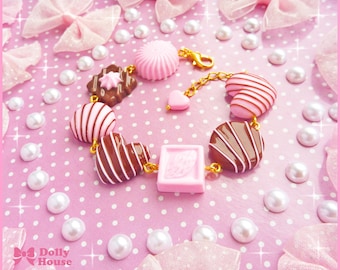 May include: A charm bracelet featuring miniature candy-themed charms including brown and pink chocolate hearts, a pink peppermint swirl, a square pink block, and a brown chocolate square with a pink star, all connected by a gold-toned metal chain.