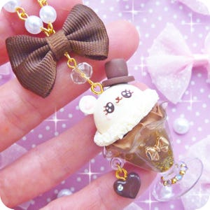 May include: A brown and white ice cream charm necklace with a small brown bow, a white ice cream cone with a brown chocolate topping, and a small brown hat on top. The charm is hanging from a gold chain with white pearl beads.