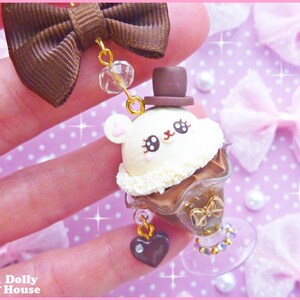 May include: A white and brown chocolate dessert charm with a cute cartoon face wearing a brown hat. The charm is attached to a brown bow and a gold chain with a heart pendant.