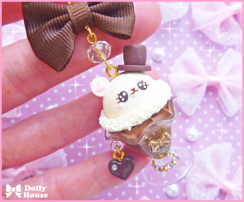 May include: A white and brown chocolate dessert charm with a cute cartoon face wearing a brown hat. The charm is attached to a brown bow and a gold chain with a heart pendant.