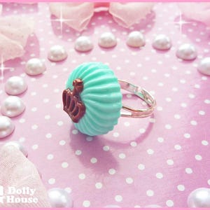 K&ouml;nnte beinhalten: Ein t&uuml;rkisblauer Cupcake-Ring mit einem braunen Kronendesign. Der Ring befindet sich auf einem rosa Hintergrund mit wei&szlig;en Punkten.