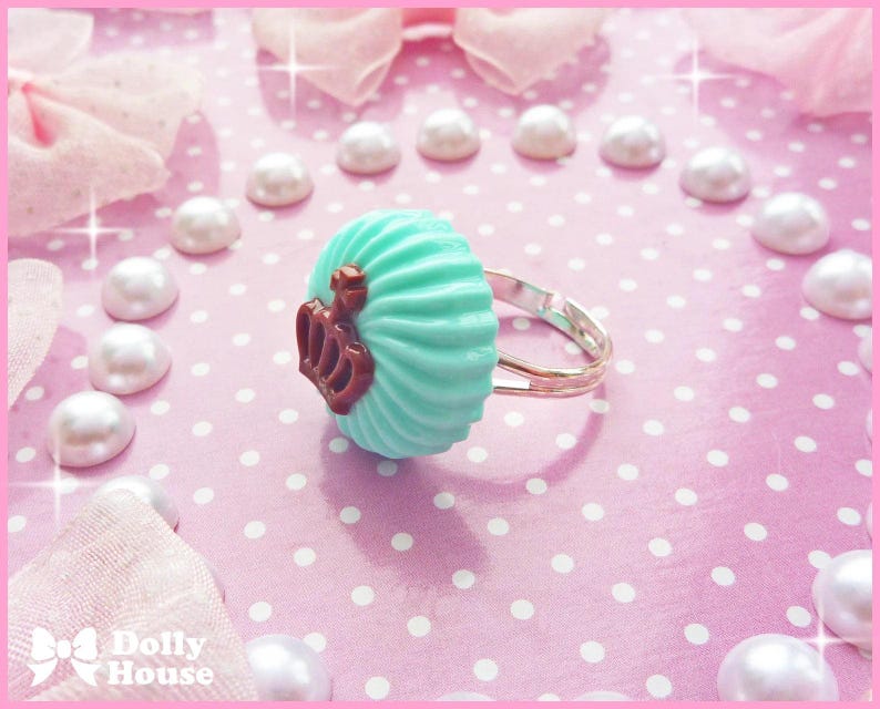 K&ouml;nnte beinhalten: Ein t&uuml;rkisblauer Cupcake-Ring mit einem braunen Kronendesign. Der Ring befindet sich auf einem rosa Hintergrund mit wei&szlig;en Punkten.
