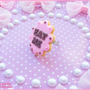May include: A pink and brown cookie ring with the words "Eat Me" written in brown icing. The ring is surrounded by white pearls on a pink background.