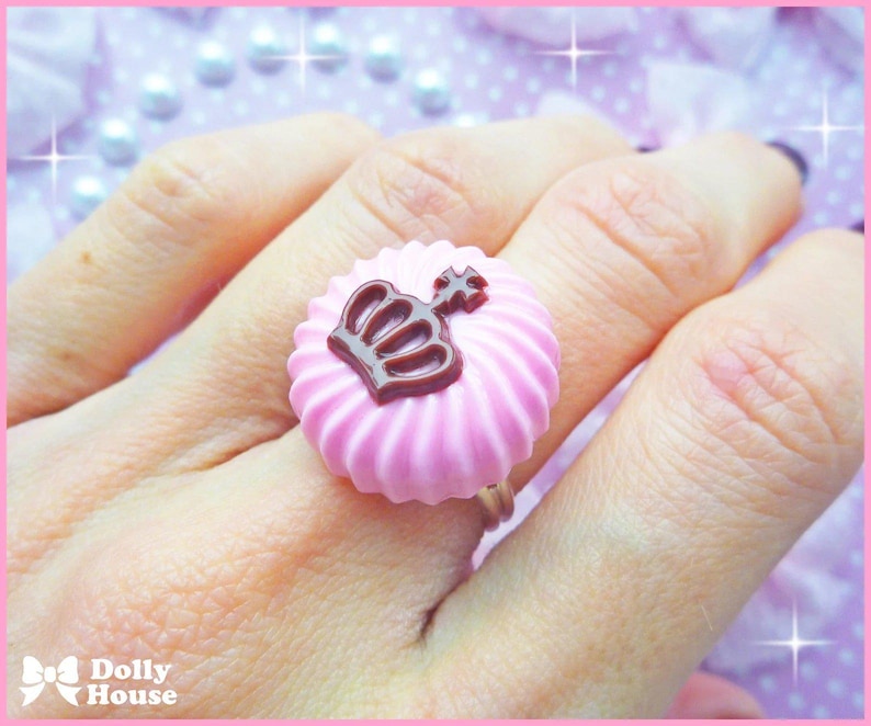 K&ouml;nnte beinhalten: Rosa Ring mit einem braunen Kronendesign. Der Ring ist in Form eines Zuckerguss-Wirbels geformt.