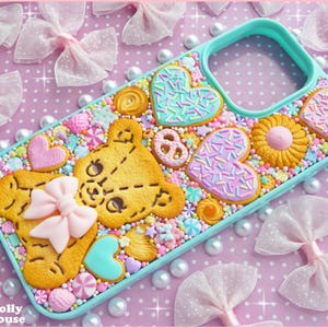 Op de afbeelding: Een turquoise telefoonhoesje versierd met een verscheidenheid aan kleurrijke, eetbaar uitziende snoepjes. Het ontwerp omvat een teddybeer, harten en bloemvormige koekjes, allemaal versierd met hagelslag en een roze strik. De achtergrond is een roze oppervlak met stippen.