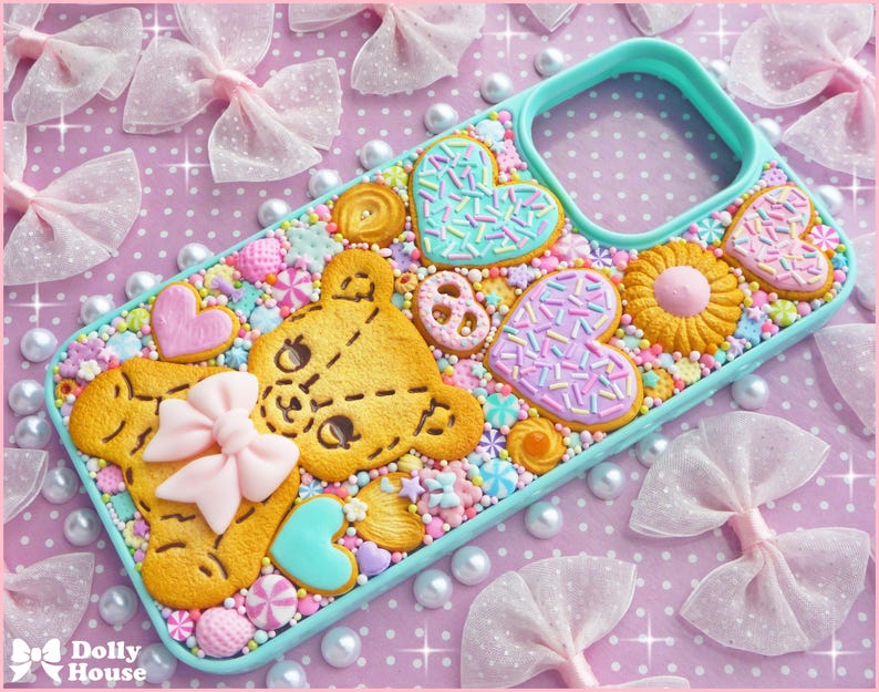 Op de afbeelding: Een turquoise telefoonhoesje versierd met een verscheidenheid aan kleurrijke, eetbaar uitziende snoepjes. Het ontwerp omvat een teddybeer, harten en bloemvormige koekjes, allemaal versierd met hagelslag en een roze strik. De achtergrond is een roze oppervlak met stippen.