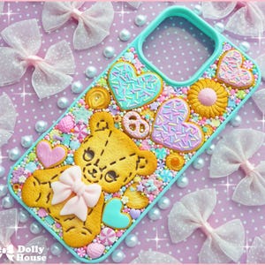 Op de afbeelding: Een turquoise telefoonhoesje versierd met een teddybeerkoekje, hartvormige koekjes en andere bakwaren. De hoes is versierd met kleurrijke hagelslag en een roze strik. De achtergrond heeft witte strikken en parels.