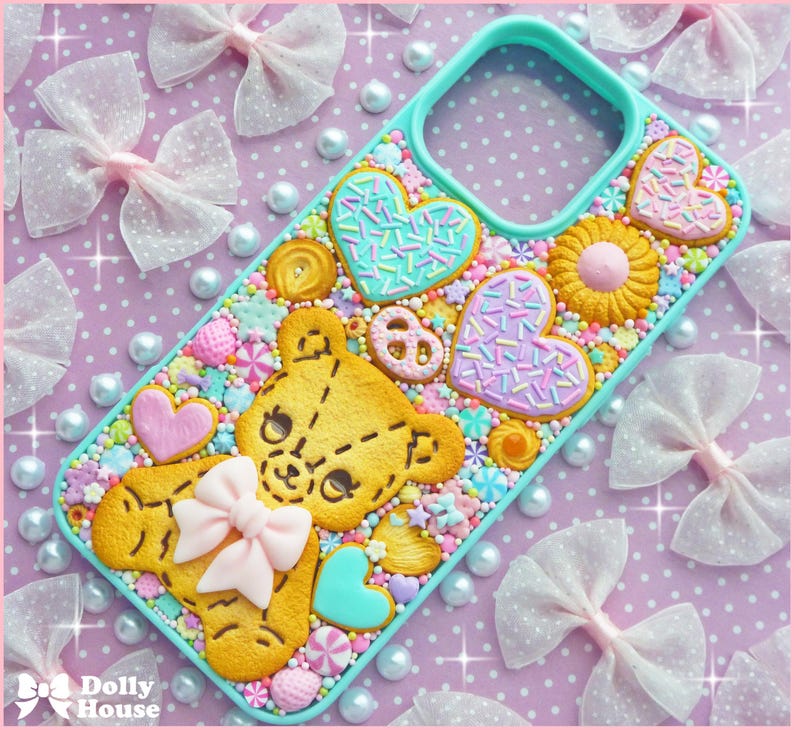 Op de afbeelding: Een turquoise telefoonhoesje versierd met een teddybeerkoekje, hartvormige koekjes en andere bakwaren. De hoes is versierd met kleurrijke hagelslag en een roze strik. De achtergrond heeft witte strikken en parels.