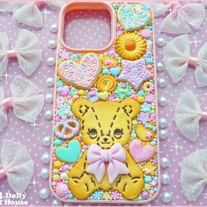 Op de afbeelding: Een roze telefoonhoesje versierd met een gele teddybeer, hartvormige koekjes en snoepjes. De hoes heeft een strik en wordt omringd door roze strikken en parels op een roze stippenachtergrond.