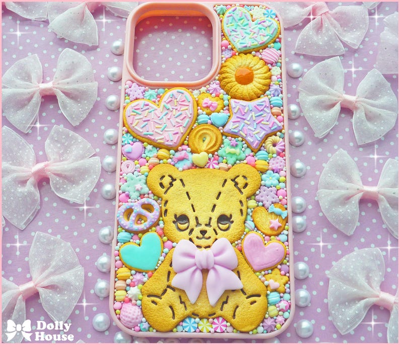 Op de afbeelding: Een roze telefoonhoesje versierd met een gele teddybeer, hartvormige koekjes en snoepjes. De hoes heeft een strik en wordt omringd door roze strikken en parels op een roze stippenachtergrond.