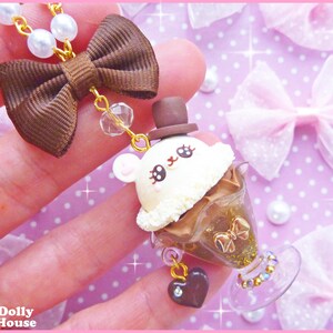 May include: A brown bow with a gold chain and a white and brown ice cream charm with a bunny face. The charm is in a clear glass container with gold glitter and a heart-shaped charm.