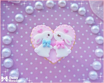Op de afbeelding: Een roze hartvormige koekje versierd met twee witte konijntjes, een met een blauwe en een met een roze strik. Het koekje ligt op een roze achtergrond met witte stippen.