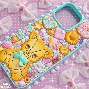 Op de afbeelding: Een decoratieve telefoonhoes met een geel teddybeerontwerp met een blauwe strik, omringd door kleurrijke snoepjes en koekjes. De hoes heeft een turquoise rand en staat op een roze achtergrond met stippen, strikken en parels.