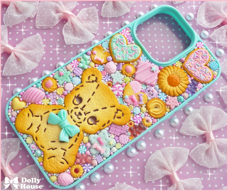 Op de afbeelding: Een decoratieve telefoonhoes met een geel teddybeerontwerp met een blauwe strik, omringd door kleurrijke snoepjes en koekjes. De hoes heeft een turquoise rand en staat op een roze achtergrond met stippen, strikken en parels.