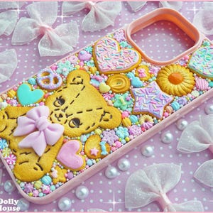 Op de afbeelding: Een roze telefoonhoesje versierd met een teddybeerkoekje, hartvormige koekjes en stervormige koekjes, allemaal versierd met hagelslag. De hoes is versierd met kleurrijke snoepjes en een roze strik. De achtergrond heeft witte strikken en parels.