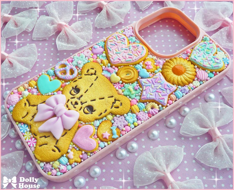Op de afbeelding: Een roze telefoonhoesje versierd met een teddybeerkoekje, hartvormige koekjes en stervormige koekjes, allemaal versierd met hagelslag. De hoes is versierd met kleurrijke snoepjes en een roze strik. De achtergrond heeft witte strikken en parels.