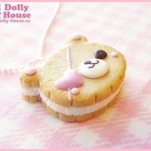 Puede incluir: Una galleta marr&oacute;n y blanca con forma de oso con un coraz&oacute;n rosa y una cara blanca. La galleta tiene un relleno blanco y est&aacute; sobre un fondo rosa y blanco.