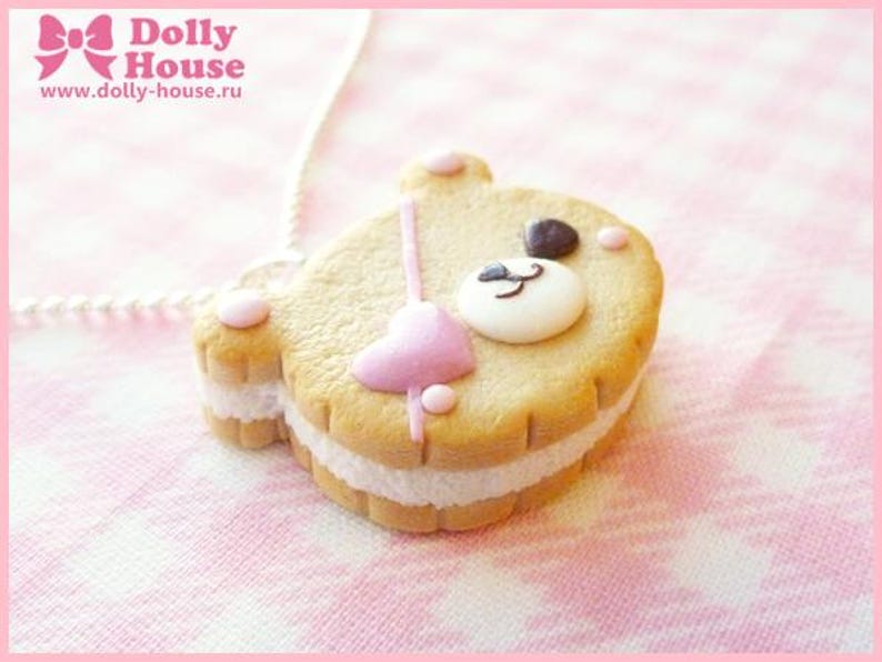 Puede incluir: Una galleta marr&oacute;n y blanca con forma de oso con un coraz&oacute;n rosa y una cara blanca. La galleta tiene un relleno blanco y est&aacute; sobre un fondo rosa y blanco.