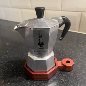 May include: A silver Bialetti Moka Express coffee maker with a black handle and lid, sitting on a wooden stand. The coffee maker has the Bialetti logo and the text "Moka Express Made in Italy".