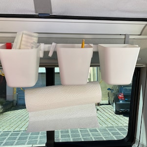 Könnte beinhalten: Drei weiße Plastikbehälter, die an einer Metallstange hängen, mit einer Rolle Papierhandtücher, die darunter hängen. Die Behälter sind leer, aber einer enthält einen gelben Bleistift. Die Behälter sind in einem Fahrzeug montiert.