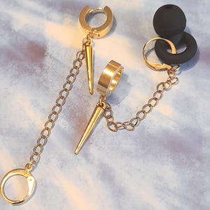 May include: Gold-toned earrings with a chain and spike design. One earring features a hoop and a chain that connects to a spike and a small hoop. The other earring has a black rubber plug with a gold hoop and chain.