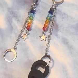 May include: A pair of dangle earrings with a rainbow bead design. Each earring features a chain, a star charm, and a black, rounded shape at the bottom. The earrings have silver-colored ear wires.