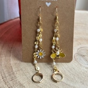Peut inclure: Une paire de boucles d'oreilles pendantes dorées avec un charme d'abeille et de marguerite. Les boucles d'oreilles sont faites avec une chaîne de perles dorées et de perles blanches.