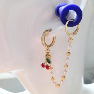 May include: Gold-tone earrings with a cherry charm and a pearl chain. One earring is a small hoop with clear stones, and the other is a larger hoop with a chain of pearls and gold beads. A blue ear gauge is visible.