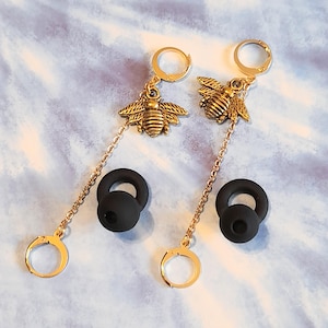 May include: Gold-colored bee earrings with black earbud attachments. The earrings feature a detailed bee charm, a delicate chain, and a small hoop. The background is a light blue and white marbled pattern.
