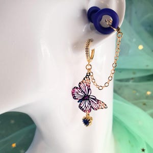 May include: A gold-toned earring with a butterfly pendant featuring pink, purple, and white details. The earring includes a small blue heart-shaped charm and a chain connecting to a blue ear plug. The earring is on a white surface.
