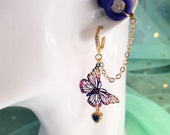 May include: A gold-toned earring with a butterfly pendant featuring pink, purple, and white details. The earring includes a small blue heart-shaped charm and a chain connecting to a blue ear plug. The earring is on a white surface.