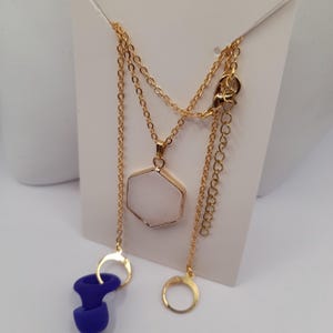 May include: A gold chain necklace with a white hexagonal pendant. The necklace is on a white card with two gold rings and a pair of blue earplugs.