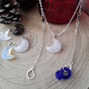 May include: A silver chain necklace with a crescent moon pendant made of white gemstone. The necklace is layered with a second silver chain with star charms. The necklace is displayed on a wooden surface with other crescent moon pendants.