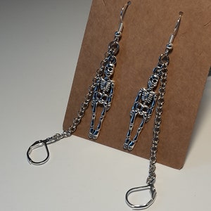 May include: A pair of silver-colored skeleton earrings. Each earring features a detailed skeleton charm hanging from a chain, with a lever-back closure. The earrings are displayed against a neutral background.