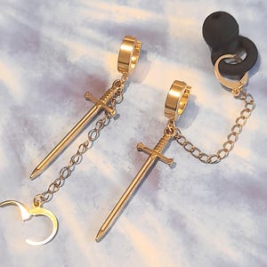 May include: Gold-colored sword earrings with a chain and a black ear cuff. The earrings feature a sword pendant and a hoop design. The chain connects the sword to the ear cuff.