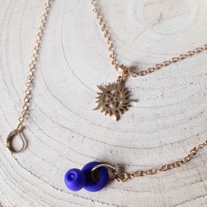 May include: Gold-coloured necklace with a sunburst pendant featuring a pentagram design. The necklace is paired with a gold chain and a blue earring with a ring and a small ball. The jewellery is displayed on a light wood surface.