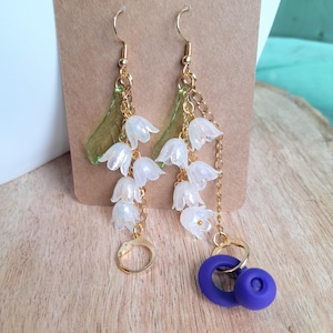 May include: A pair of dangle earrings featuring white flower clusters, gold chains, and a purple accent. The earrings have gold-colored hooks and are set against a neutral background. The earrings are designed to resemble lily of the valley flowers.