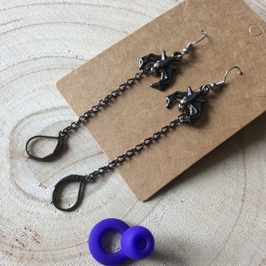 May include: A pair of long silver chain earrings with a bat charm hanging from each chain. The earrings are on a brown background.