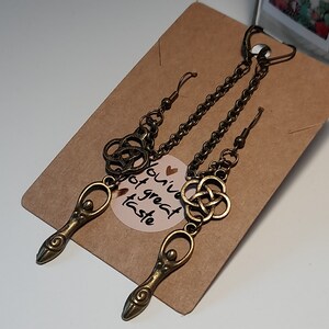 May include: A pair of antique bronze-toned earrings. Each earring features a chain, a Celtic knot, and a stylized goddess pendant. The earrings are displayed on a brown card with a sticker that reads "You've got great taste."