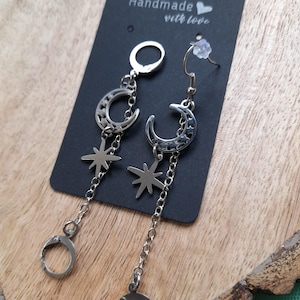 May include: Silver-toned dangle earrings featuring crescent moons and star charms. The earrings hang from a chain and have a lever-back closure. The earrings are displayed on a black card with the words "Handmade with love".