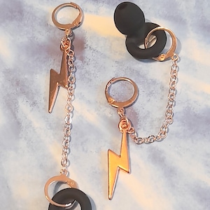 May include: Rose gold lightning bolt earrings with a black rubber ear cuff. The earrings feature a lever back closure and a chain that connects the lightning bolt to the ear cuff. The earrings are displayed on a blue and white background.