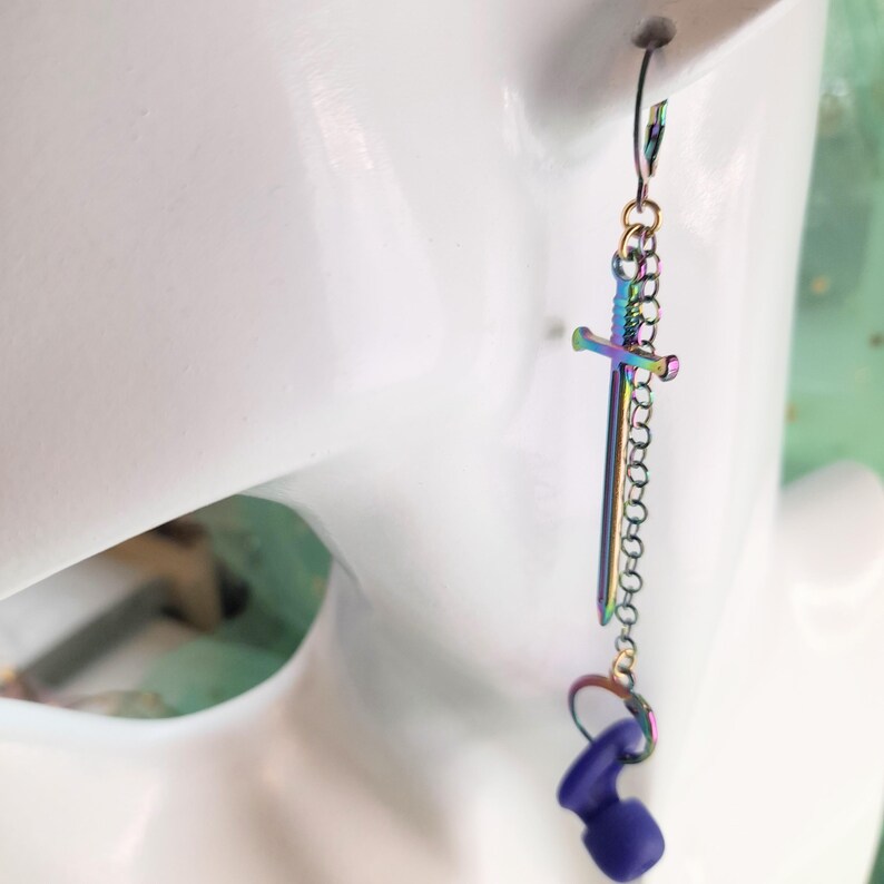 May include: A single, iridescent sword-shaped earring with a chain and a small, purple, rounded charm. The earring is suspended from a hook and is shown against a white background.
