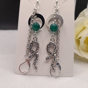 May include: Silver-coloured drop earrings with a crescent moon, a green bead, and a snake charm. The earrings have a chain design and a lever-back closure. The earrings are displayed on a white surface.