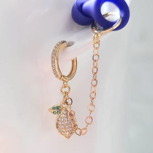 May include: Gold-toned hoop earring with a dangling lemon charm. The hoop is encrusted with small clear stones. The lemon charm is detailed with clear and green stones, and is connected to a chain that attaches to a blue object.