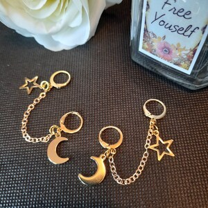 May include: Three pairs of gold hoop earrings with dangling charms. Each pair has a hoop earring with a chain attached to a charm. The charms are a crescent moon, a star, and another crescent moon.