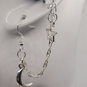May include: Silver chain earrings with a crescent moon and star charm. The earrings are connected by a chain.