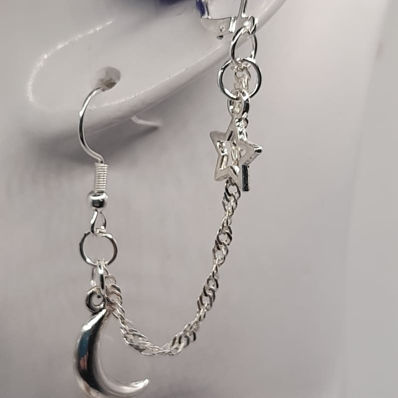 May include: Silver chain earrings with a crescent moon and star charm. The earrings are connected by a chain.